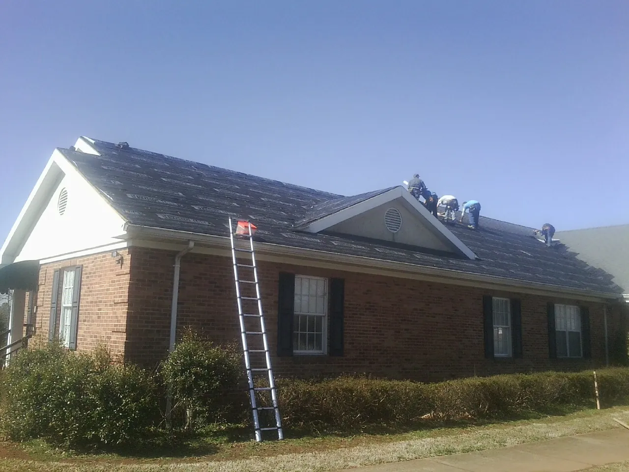 Crew tearing shingles off a brick ranch roof with a ladder set — Emergency Roof Repair work by our crew in Guin