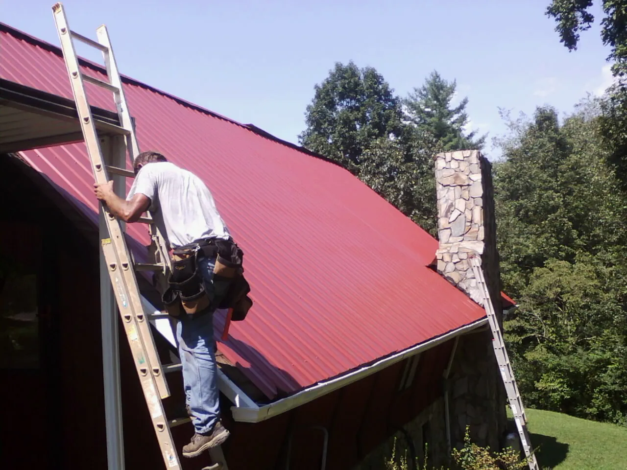 Roofer on a ladder beside a red metal roof and stone chimney — Soffit & Fascia Repair by Roofing Brothers in Guin